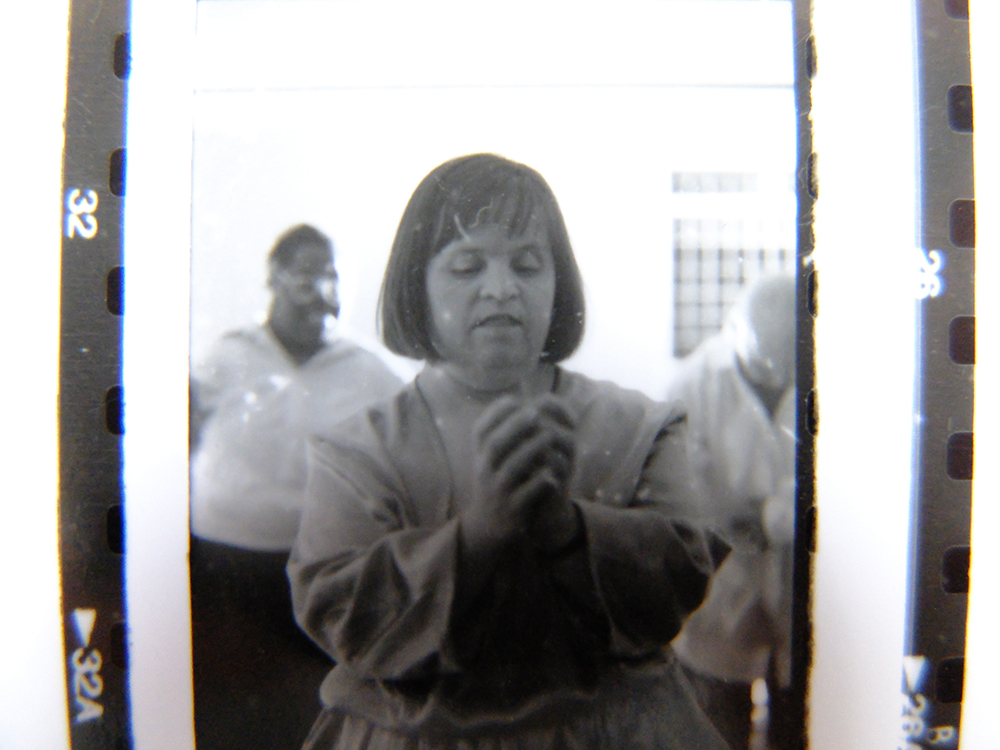A black & white photo still of the performance Diary of a Dead Artist  by Corali Dance Company. The photo is taken in the original gallery of Southwark Park Galleries and features Margaret Courtman with her hands cupped around each other in a gesture. She wears an old fashioned outfit.
